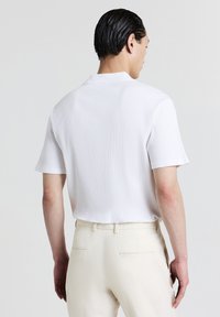 Camicia bianca a righe con maniche corte e colletto polo, vista da dietro, abbinata a pantaloni chiari con un motivo testurizzato.