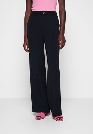 Femme portant un pantalon noir à jambes larges, un haut rose sans manches et des sandales roses à talons, debout devant un fond blanc uni.