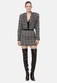 Kobieta w szarej kratkowanej marynarce o kroju cropped i pasującej mini spódnicy, w czarnych skórzanych kozakach za kolano, stojąca na białym tle.