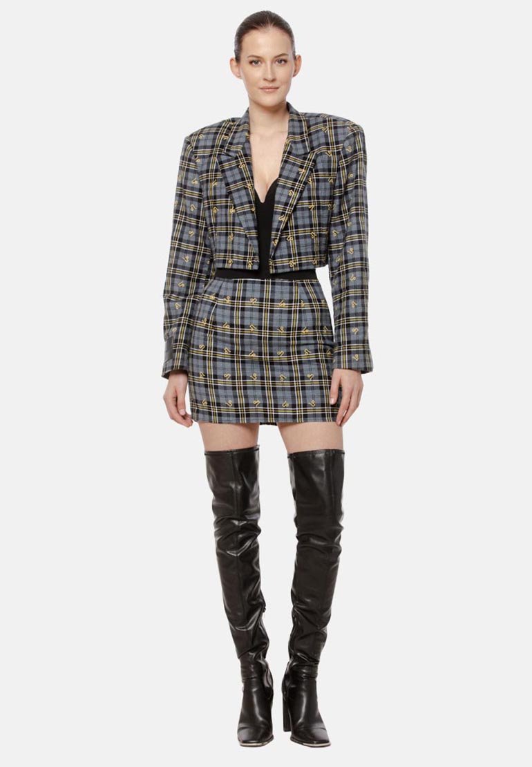 Kobieta w szarej kratkowanej marynarce o kroju cropped i pasującej mini spódnicy, w czarnych skórzanych kozakach za kolano, stojąca na białym tle.