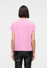 Γυναίκα μ ε κοντά σκουρόχρωμα μαλλιά, φοράει υφασμάτινο ανοιχτό ροζ κοντομάνικο τοπ και μαύρο δερμάτινο παντελόνι, εμφανίζεται από πίσω σε απλό φόντο.