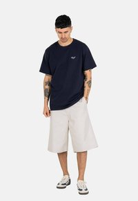 Navy blue short-sleeve t-shirt with a relaxed fit and a small graphic on the chest, paired with light beige knee-length shorts and sneakers.