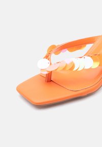 Πορτοκαλί σανδάλι με τετράγωνη μύτη και λεπτό λουράκι που διακοσμείται με μεγάλα, λαμπερά, στρογγυλά παγιέτες σε ανοιχτό φόντο.