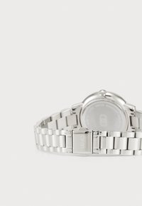 Zilveren metalen horloge met een schakelpand, ronde kast en gegraveerde sluiting; heeft een doorzichtige achterkant die de interne mechanismen toont.