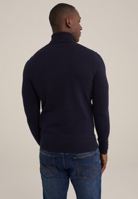 Tengerészt navy kötött garbó pulóver hosszú ujjal és bordás mandzsettával. Kék farmernadrággal párosítva, amelyen kontrasztos varrás található a zsebeknél.