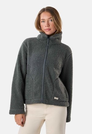 Ung kvinde iført en mørkegrå fleecejakke med lynlås og lyse beige bukser, stående mod en ensfarvet lys baggrund.