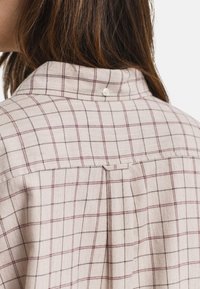 Mulher com cabelo castanho a usar uma camisa às riscas beige com linhas vermelhas e pretas, mostrando os detalhes da gola e dos ombros nas costas.
