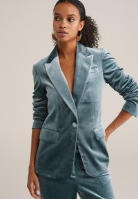 Sammetsblazer i teal, med en enkel knapp, spetsiga slag och framfickor. Slät struktur med formade ärmar, stylad öppen.