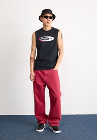 Juodas be rankovių Reebok marškinėliai su logotipu, raudoni džinsai, juodas kibiro skrybėlė ir saulės akiniai, derinami su juodais ir baltais sportbačiais.