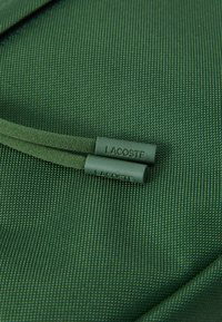 Zeleni teksturirani materijal s dva povlačna užeta koja imaju zelene plastične vrhove s natpisom "LACOSTE". Usredotočeno na detalje materijala i dizajna.