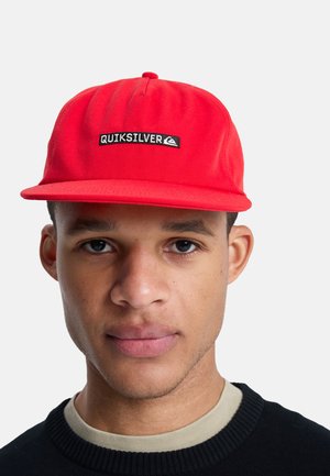 Young man wearing a bright red Quiksilver cap and a black sweater with light beige collar against a plain white background.