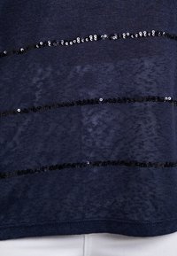 Top azul marino con franjas horizontales de lentejuelas negras sobre una tela texturizada. El diseño tiene un corte relajado y un sencillo escote redondo.