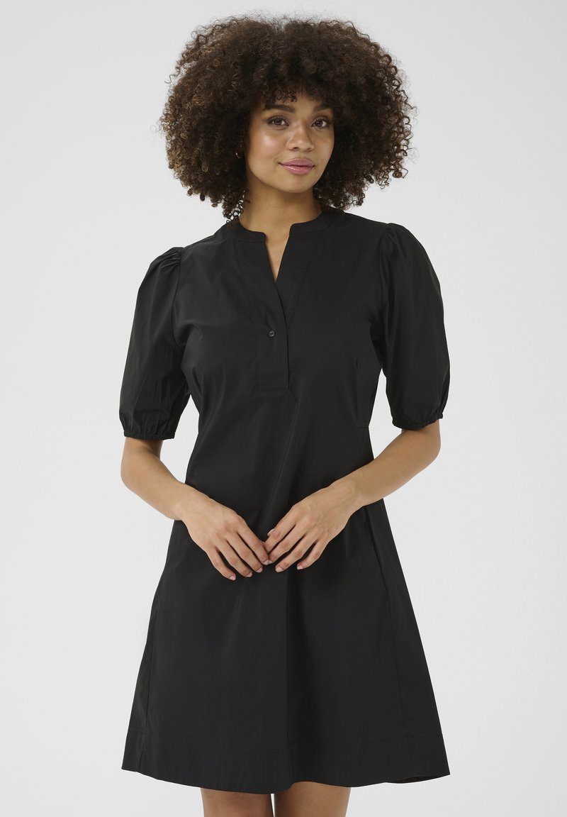 Crna kratka haljina s kratkim rukavima i stojećim ovratnikom, s detaljima na gumbima i opuštenim krojem. Izrađena od glatkog materijala s blago teksturiranom površinom.