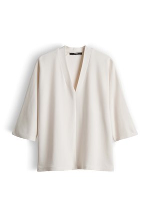 Blouse à manches longues couleur crème, col en V, coupe ample et design simple, présentée sur un fond blanc.