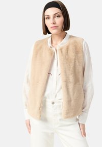 Beige fuskpälsväst med rund halsringning och utan knäppningar, bärs över en vit skjorta med knappar. Slät textur, avslappnad passform.