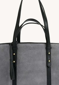 Sac fourre-tout en suede gris avec des poignées en cuir noir, des sangles réglables ornées de clous dorés, et un détail en frange.