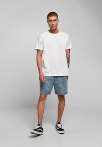 Camiseta blanca de algodón; pantalones cortos de mezclilla azul claros; zapatillas de deporte en blanco y negro; pelo corto; pose de pie frente a un fondo gris.