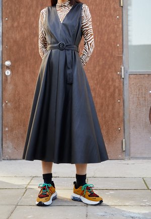 Vestido de cuero sintético negro con escote en V y cinturilla, combinado con zapatillas coloridas con cordones verdes y detalles estampados.