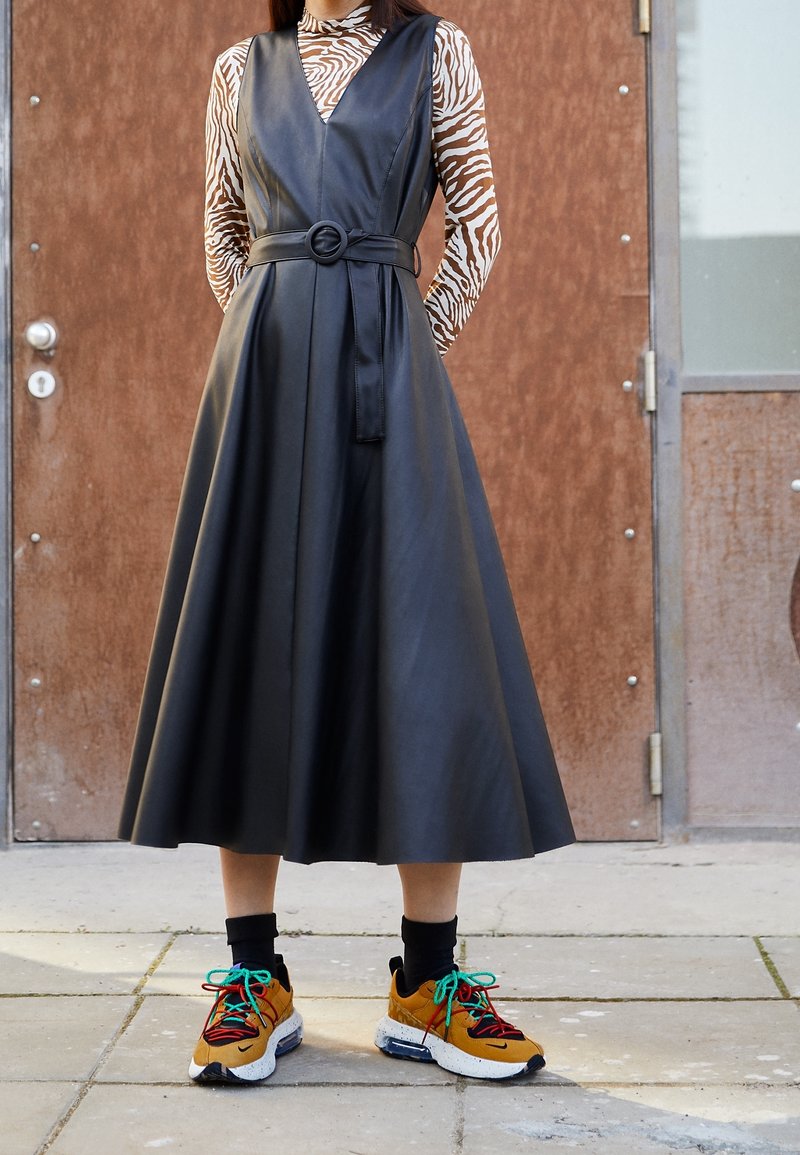 Svart faux läderklänning med V-ringning och bälte i midjan, tillsammans med färgglada sneakers med gröna snören och mönstrad detaljer.