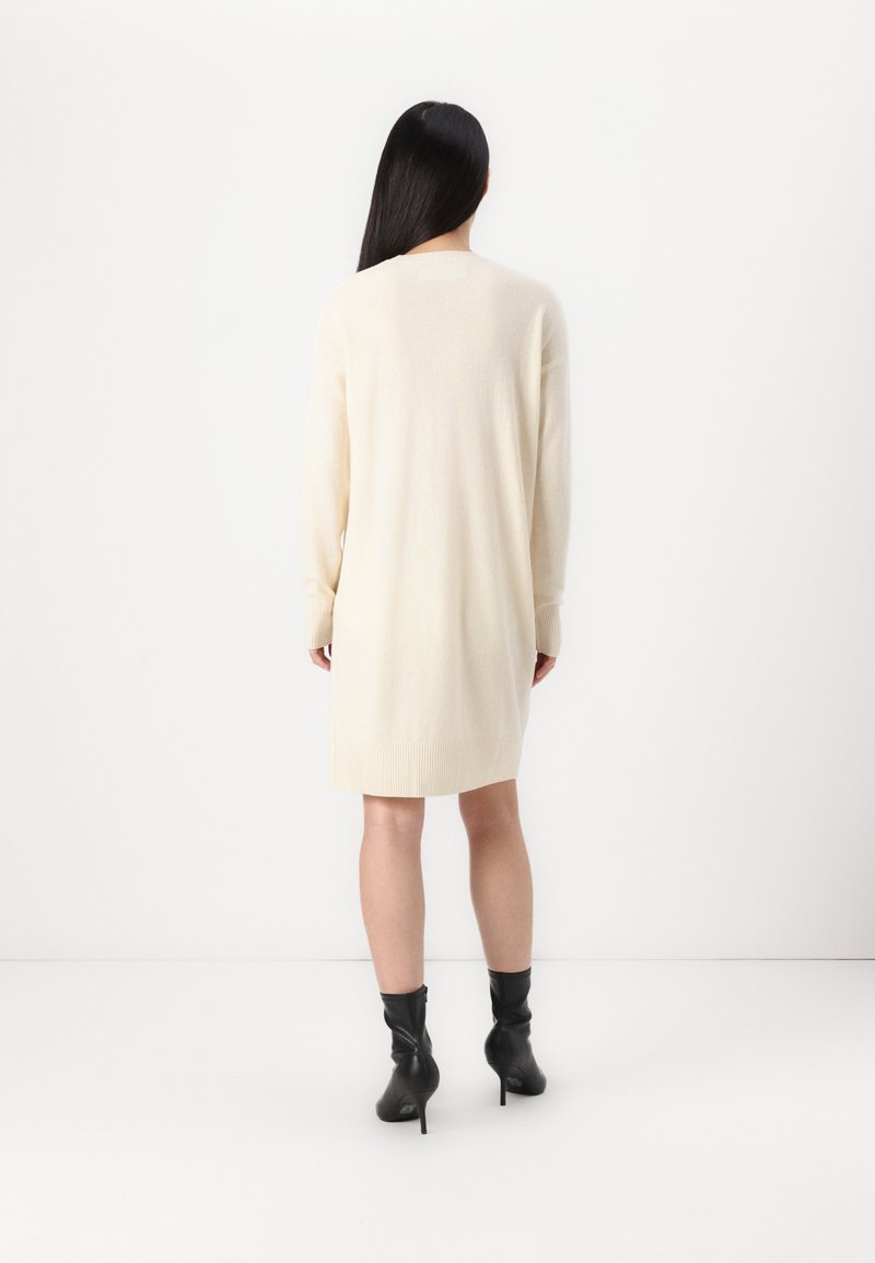 Beżowa dzianinowa sukienka z długimi rękawami, okrągłym dekoltem i ściągaczami przy mankietach, sięgająca powyżej kolan, w zestawieniu z czarnymi botkami za kostkę.