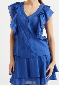 Niebieska bluzka w paski z falbaniastymi rękawami, dekoltem w serek oraz guzikami z przodu. Wykonana z lekkiego materiału, w połączeniu ze spódnicą w kształcie dzwonu w tym samym kolorze.