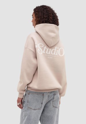 Donna con capelli ricci che indossa una felpa beige con cappuccio con la scritta "Studio" sulla schiena e jeans a vita alta color azzurro chiaro, su uno sfondo semplice.