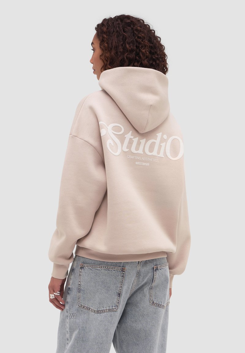 Femme aux cheveux bouclés portant un sweat à capuche beige avec le texte « Studio » au dos et un jean taille haute bleu clair, sur un fond uni.
