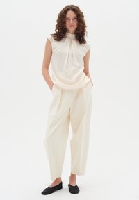 Light cream blouse with folded neckline, paired with loose-fit white trousers and black flats. Smooth fabric with minimal detailing.