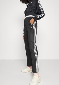 Person som bär svarta Adidas träningsbyxor och en matchande jacka med vita ränder, stående med en hand i fickan, vita sneakers.