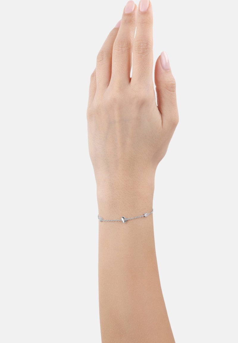 Mano con pulsera de cadena de plata con pequeños charms en forma de corazón, dedos extendidos hacia arriba contra un fondo claro y liso.
