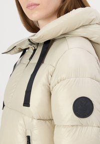 Beige puffer jacket with a hood, featuring a textured, glossy finish, black zippers, and a round logo patch on the left shoulder.