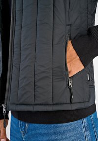 Svart vadderad väst med dragkedja, sidofickor och ribbade mudd. Materialet verkar slätt med en mjuk yta.