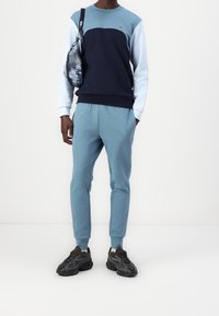 Felpa color block blu e blu scuro con maniche lunghe, abbinata a pantaloni jogger blu coordinati. Include un piccolo logo e una borsa a spalla con motivo.