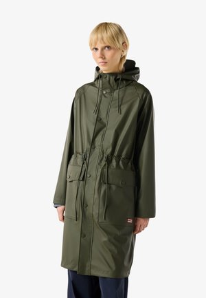 Blond osoba nosící dlouhý olivově zelený plášť s kapucí, předními kapsami a stahováním v pase, stojící na bílém pozadí.