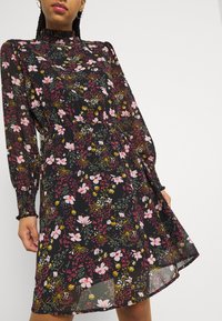 Femme portant une robe noire à motifs floraux rose, jaune et verts, manches longues en tissu transparent, et un col montant à volants.