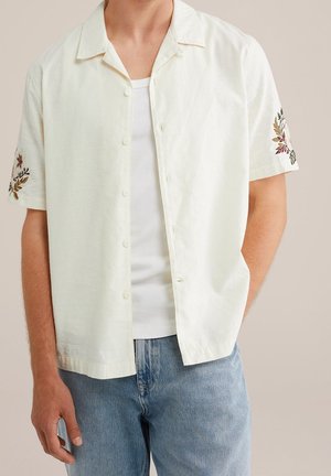Mann trägt ein cremefarbenes kurzärmeliges Hemd mit bestickten Blumendetails an den Ärmeln über einem weißen Tanktop und blauen Jeans.