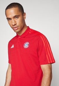 Rød polo skjorte med krave, der har tre orange striber på skuldrene og et cirkulært blåt FC Bayern München logo på brystet.