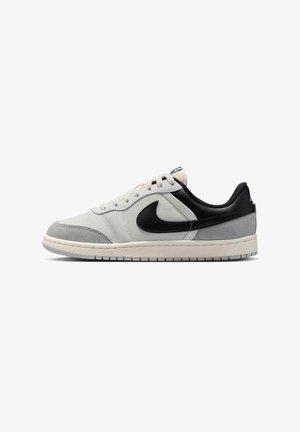 Zapatilla baja en blanco y gris con talón negro y logo de Nike, cordones blancos y suela texturizada, mostrada de lado.