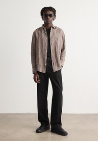 Camicia in lino marrone chiaro con chiusura a bottoni, t-shirt nera sottostante, pantaloni neri e sneakers nere, accessoriata con occhiali da sole scuri.