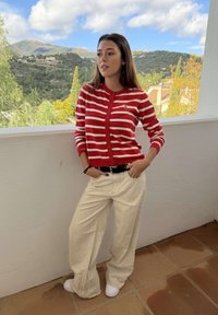 Jonge vrouw in een rood-wit gestreepte trui en beige broek met wijde pijpen staat op een balkon met een bergachtig landschap op de achtergrond.