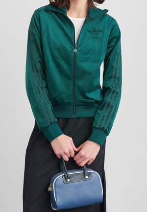 Osoba nosząca zieloną kurtkę na zamek Adidas, trzymającą małą niebieską torebkę z ciemnymi uchwytami i złotymi detalami zamka.