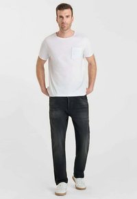 T-shirt blanc à manches courtes avec une poche, associé à un jean foncé. Le mannequin porte des baskets blanches avec des semelles beiges. Fond uni.