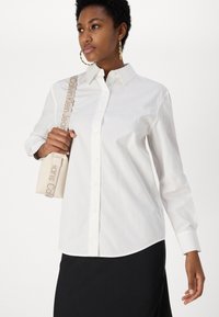 Cămașă albă cu mâneci lungi, guler și margine curbată, purtată cu o mică geantă bej, având o curea cu model.