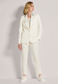 Wit pak met colbert en taps toelopende broek. Het colbert heeft een revers met grijze strepen. Het model draagt witte sneakers en staat tegen een neutrale achtergrond.