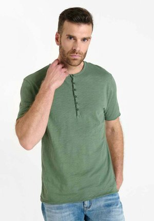 Grünes kurzärmeliges T-Shirt mit Rundhalsausschnitt und sechs Knöpfen. Der Stoff scheint weich mit einer subtilen Textur zu sein. Getragen mit hellblauen Jeans.