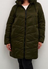 Frau trägt einen knielangen, olivgrünen gesteppte Daunenjacke mit Frontreißverschluss und Kapuze, kombiniert mit schwarzen Hosen und einem weißen Oberteil.