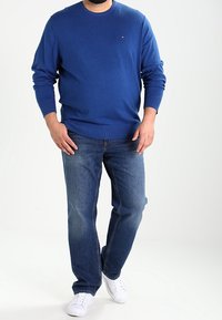 Homme portant un pull ras du cou bleu, un jean bleu et des baskets blanches, debout les mains près des poches devant un fond uni.