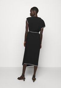 Femme portant une robe midi noire avec des bordures blanches, debout dos à la caméra sur un fond blanc uni.