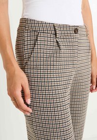 Femme portant un pantalon à carreaux marron et noir avec poches latérales et une fermeture à bouton, associé à un haut en maille blanc.