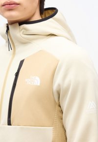 Ung person iført beige og cremefarvet North Face hættejakke med lynlås på brystlommen og Mountain Athletics-logo på ærmet.